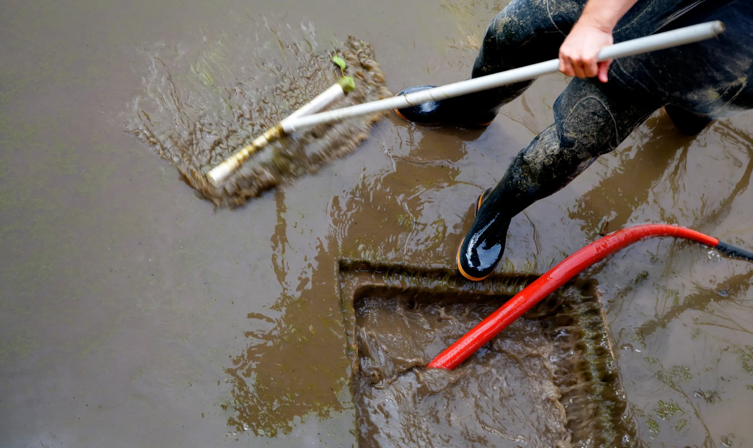 A person in rubber boots sweeping mud and floodwater into a drain.