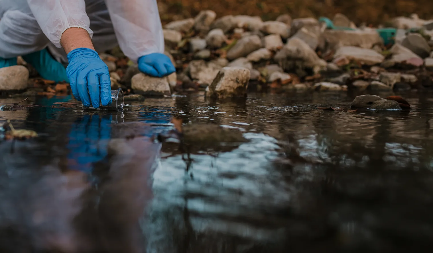 Gloved hand lowering a sample vial into a shallow stream.